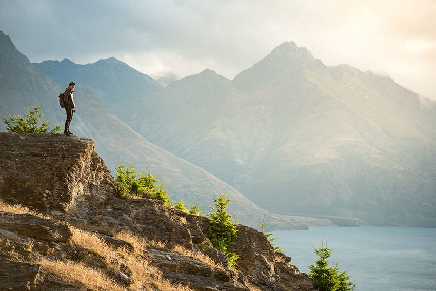 Young Asian male traveler standing on the edge of vista lookout with mountain scenery and Lake Wakatipu in background, Watching sunset in Queenstown, South Island, New Zealand