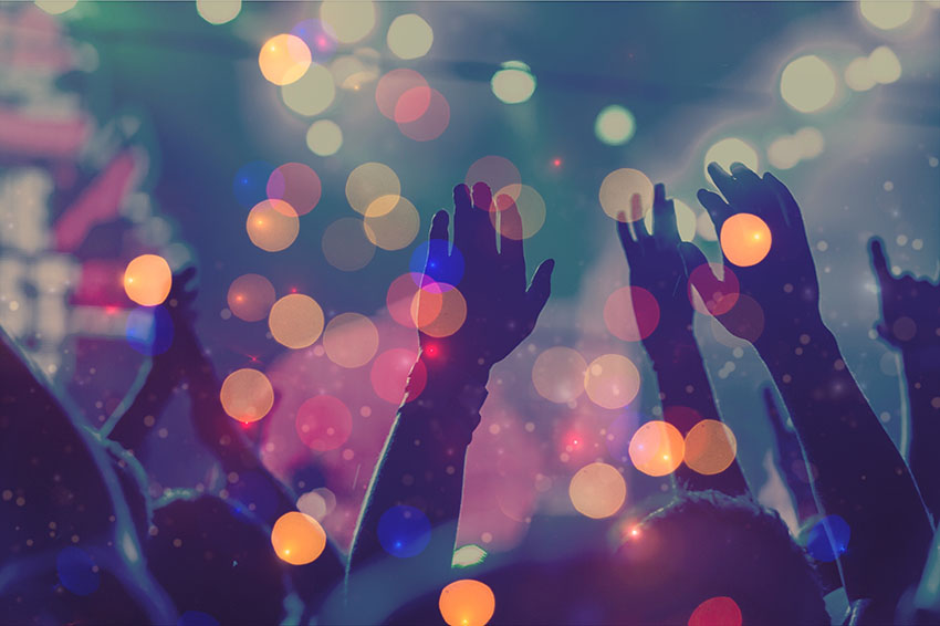 Audience with hands raised at a music festival and lights streaming down from above the stage. Soft focus, blurred movement.