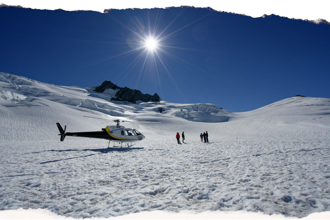 A helicopter parked on the snowfield high on a glacier, with a small group standing on the ice and crevasses running up to a rocky ridge behind