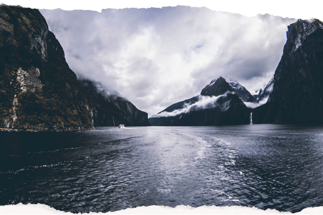 A small cruise boat under the sheer cliffs of Milford Sound, with low cloud across the peaks and waterfalls running down the rock