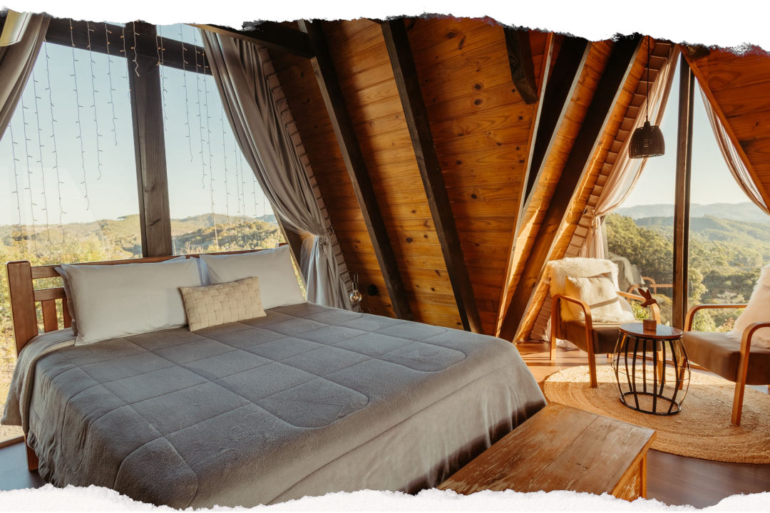 A timber A-frame lodge room in late afternoon light, with floor-to-ceiling glass on two walls looking out over wooded hills
